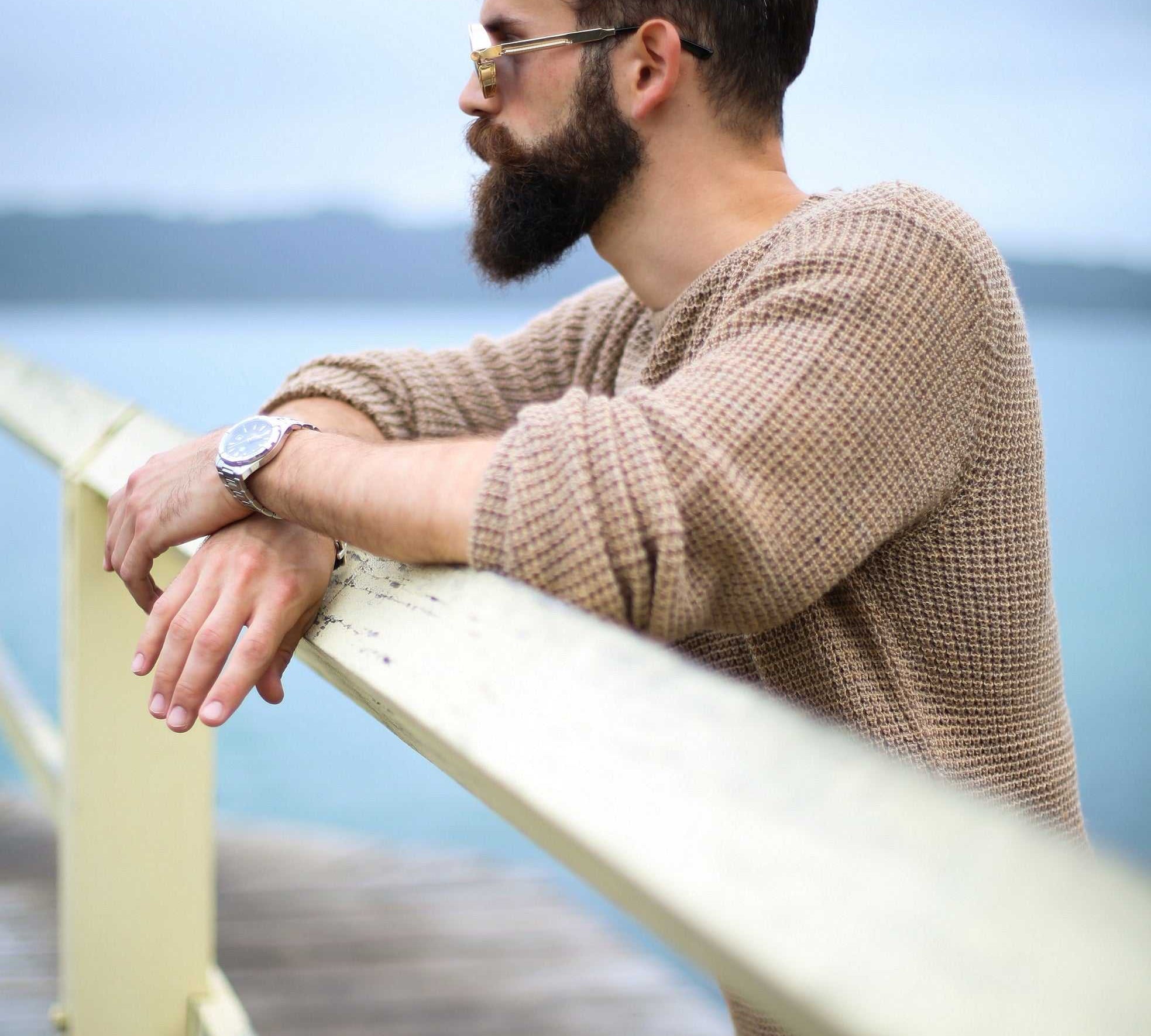 man with thick beard wearing sunglasses and brown sweater leaning on wooden railing outdoors near water
