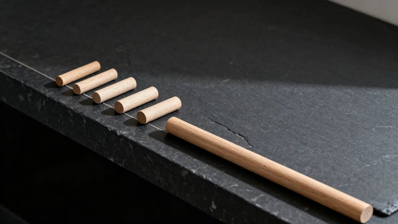 Five short oak dowels and one long oak dowel in a row on a dark slate shelf, illustrating the stages of a beard length chart