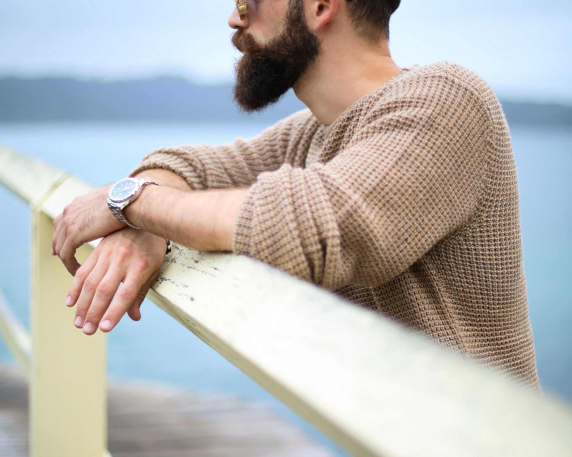 man with thick beard wearing sunglasses and brown sweater leaning on wooden railing outdoors near water
